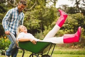 Man and woman smiling as woman is pushed along in wheelbarrow wearing pink wellington boots